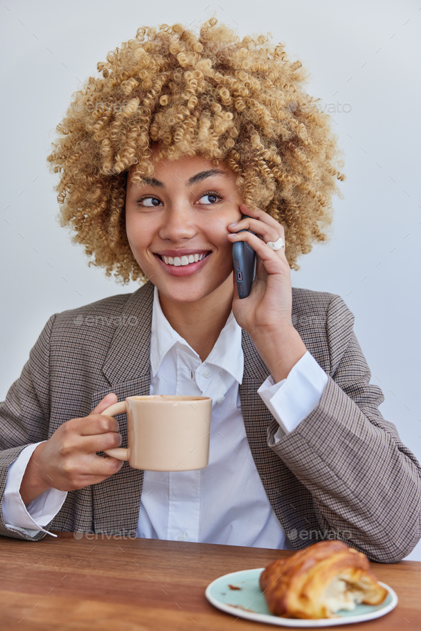 Vertical shot of smiling woman in corporate clothes makes pleasant business call sits at cozy cafete