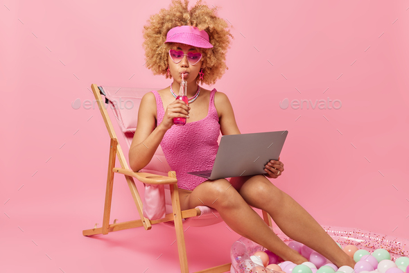 Curly haired young female model wears shades and bathing suit drinks energetic beverage watches film