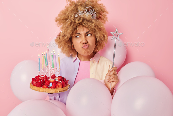 Dissatisfied curly haired young woman pouts lips looks unhappily aside holds festive cake and magic