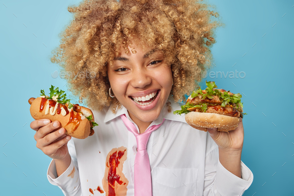 Cheat meal and junk food concept. Positive curly haired woman holds delicious hot dog and hamburger