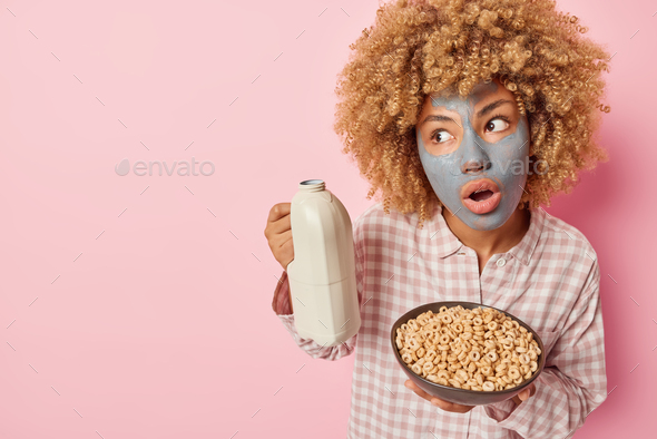 Surprised shocked woman with curly bushy hair holds breath from amazement poses with cereals and mil