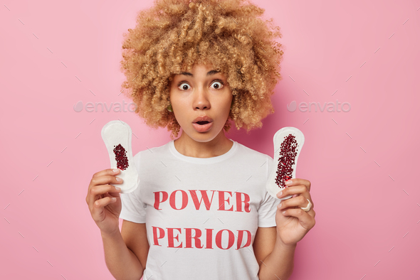 Shocked woman with curly hair holds breath from amazement holds two sanitary napkins smeared with bl
