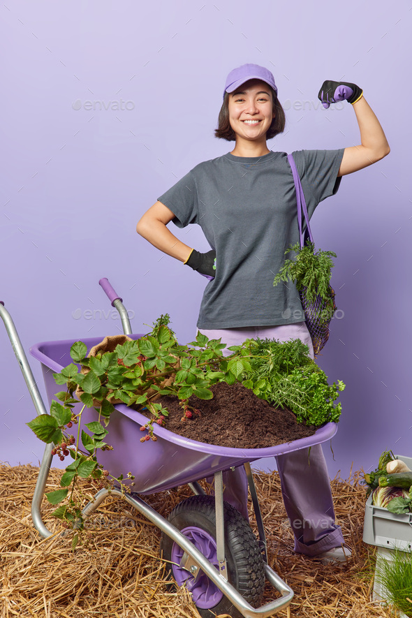 Boastful female gardener raises arm shows biceps does physical work in garden poses near homegrown v