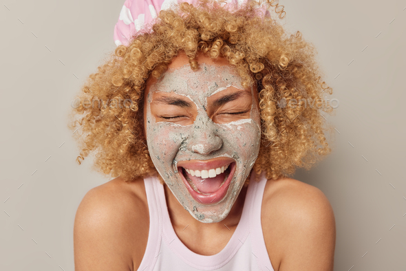 Headshot of positive curly haired woman applies beauty mud mask for reducing fine lines giggles happ