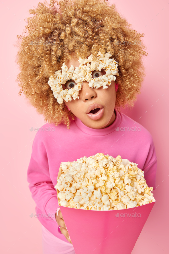 Surprised amazed woman holds big paper bucket of popcorn looks through handmade glasses pretends bei