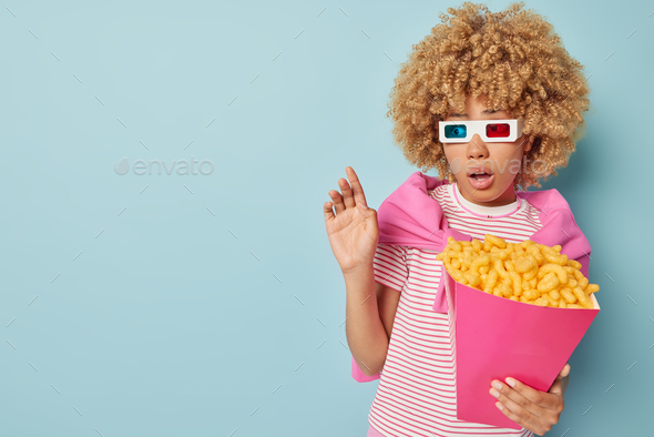Indoor shot of scared curly haired woman watches horror film in cinema poses with bucket of corn sti