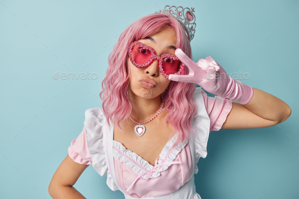 Horizontal shot of pink haired housemaid makes peace gesture over eye pouts lips wears crown sunglas
