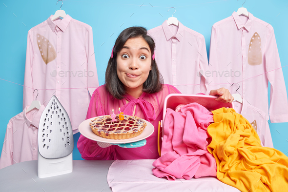 Surprised young Asian woman stares impressed licks lips holds appetizing cherry cake stands near iro