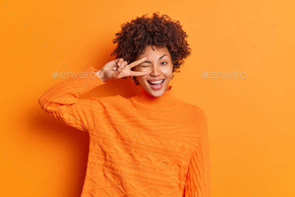 Indoor shot of happy curly haired woman makes peace gesture over eye shows v sign smiles toothily dr