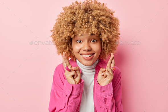 Positive hopeful woman with curly hair keeps fingers crossed smiles gladfully believes in dreams com