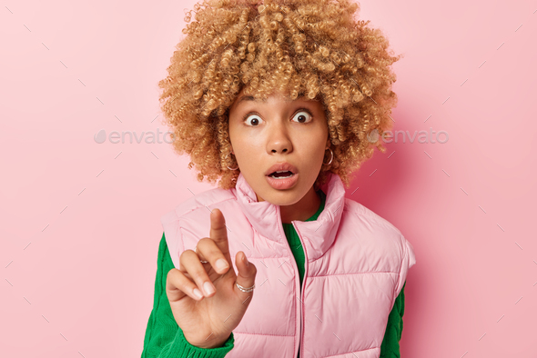 Photo of amazed curly haired young European woman keeps finger raised holds breath cannot believe ow
