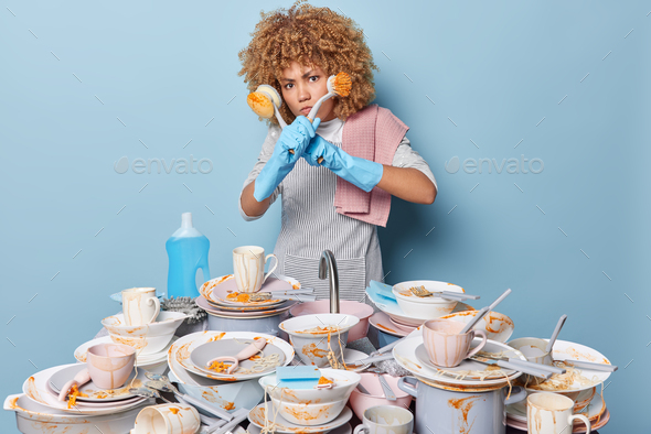 Serious housewife wears striped apron and rubber gloves surrounded by heap of dirty dishes kitchenwa