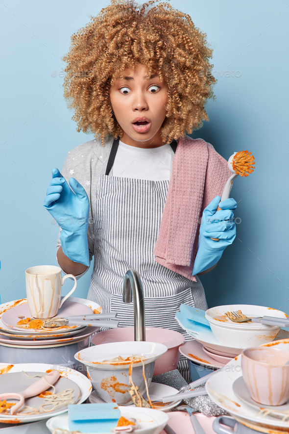 Shocked curly haired woman stares at sink full of dirty dishes holds brush wears striped apron and p