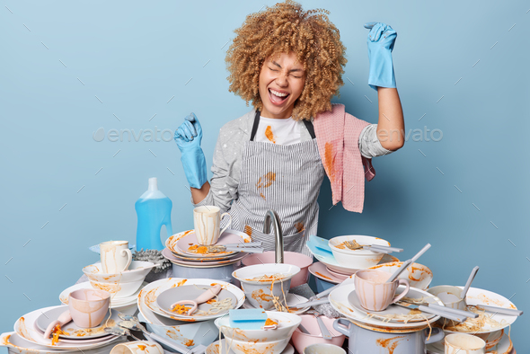 Positive housewife dances near sink full of dirty dishes with leftover food wears striped apron and