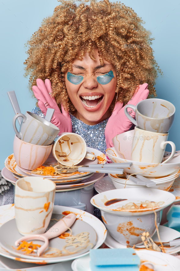 Emotional housewife poses near stack of dirty dishes keeps hands raised