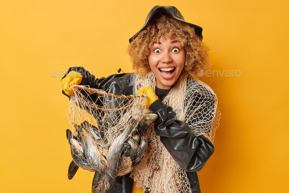 Positive surprised female angler holds fishing net full of fishing net  wears black hat and raincoat Stock Photo by wayhomestudioo