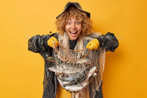 Amazed cheerful curly female fisher holds net full of big fish feels very happy to have successful f