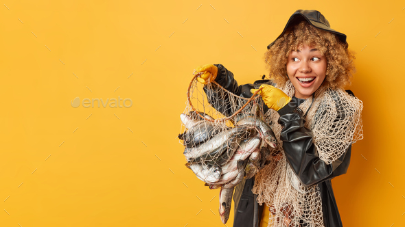 Impressed cheerful curly female fisher poses with net full of caught fish dressed in black leather h