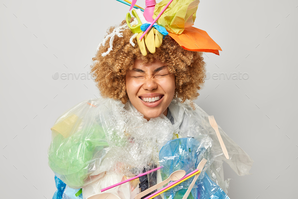 Positive curly haired female model poses with plastic garbage on head makes costume of wastes cleans