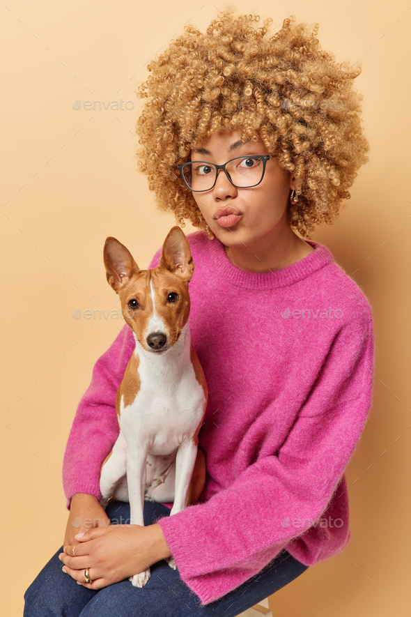 Vertical shot of curly female pet owner poses with basenji dog enjoys company of best friend pose on