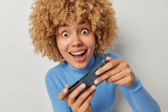 Indoor shot of amazed curly haired teenage girl reacts to super cool video game holds mobile phone s