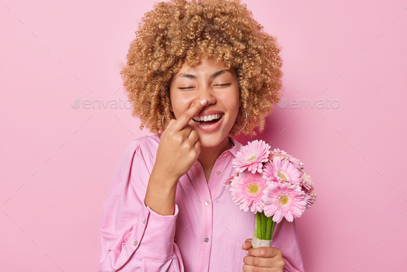 Joyful European woman with curly hair touches nose with finger keeps eyes closed has fun holds beaut