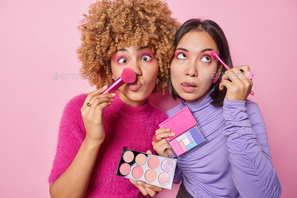 Indoor shot of two female friends holds palette of colored eye shadow use professional cosmetic brus