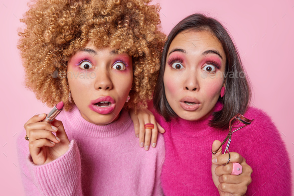 Photo of shocked mixed race women apply makeup hold lipstick and eyelashes curler prepare for party