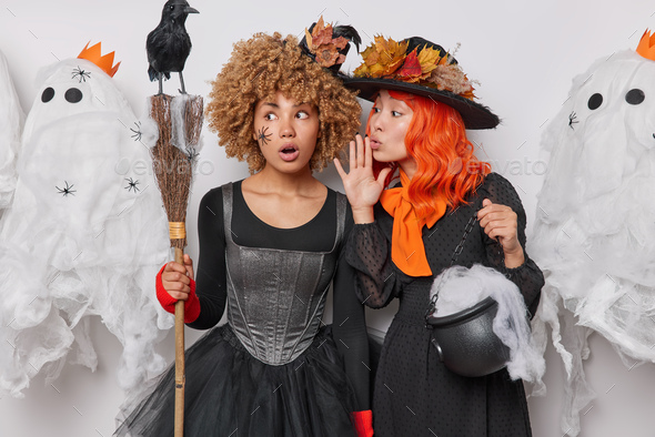 Mysterious female wizard with orange hair holds cauldron whispers secret on ear to her friend wear b