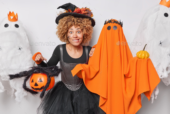 Curly haired female witch bites lips stares impressed at camera holds carved pumpkin wears black dre
