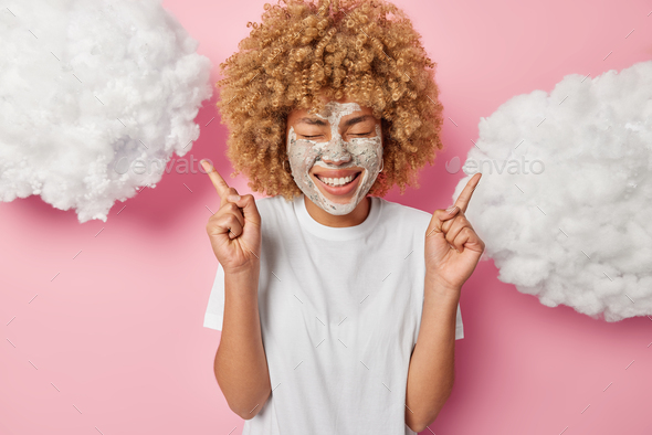 Waist up shot of positive young woman with curly hair applies beauty mask for reducing fine lines po