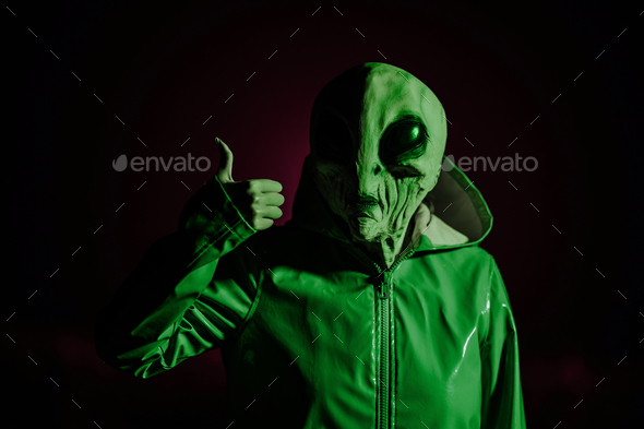 Alien showing thumb up. Like gesture. Creepy mask of humanoid on neon planet in smooth raincoat.
