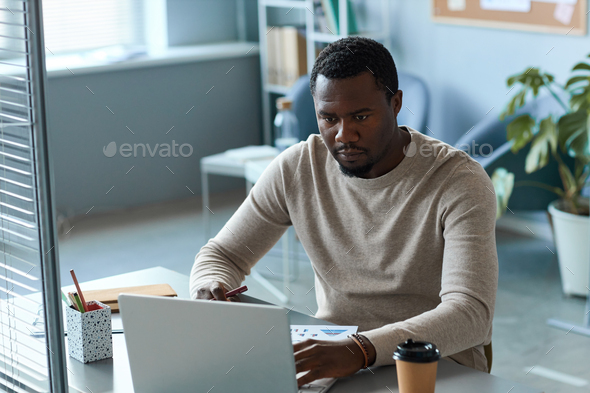 Focused black man using laptop in office behind glass partition wall ...