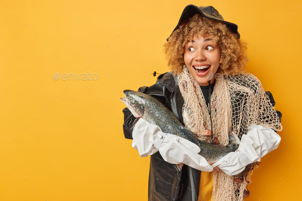 Happy surprised female fisher poses with big salmon smiles broadly wears black leather hat raincoat