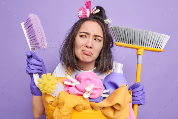 People and housework concept. Dissatisfied Asian woman holds brush and broom looks sadly aside wears