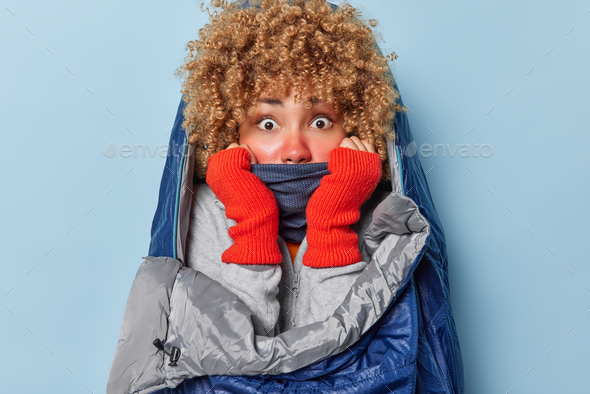 Surprised shocked curly woman hides face behind scarf wears warm gloves feels cold as sleeps
