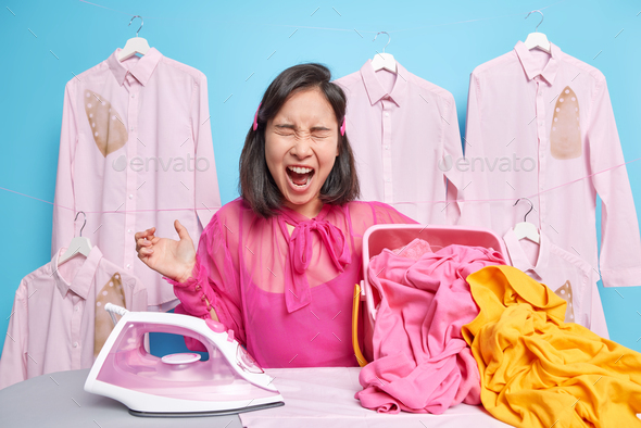 Emotional Asian woman with dark hair shouts loudly feels very angry as has to iron laundry after