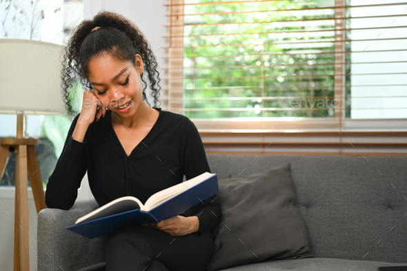 Calm young African woman reading favorite literature on couch, spending leisure time at home.