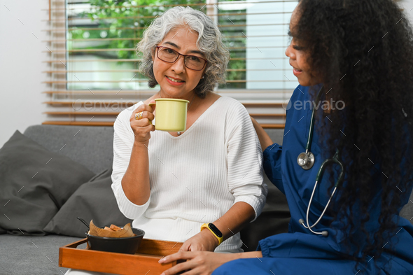 Smiling elderly woman having healthy nutrition breakfast meal with helpful caregiver on couch.