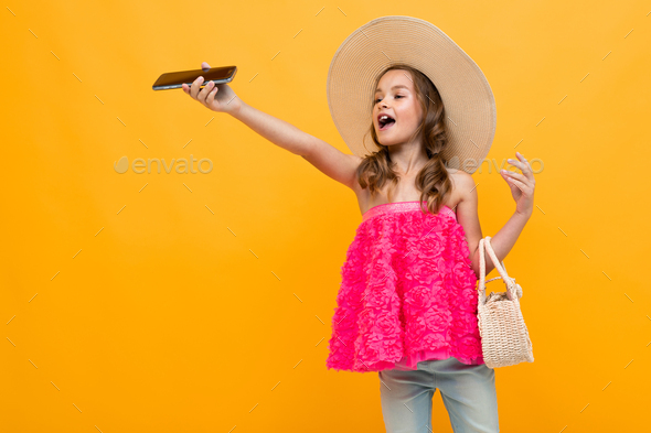 caucasian stylish girl in a straw hat with a round bag raised her hand with a phone on an orange