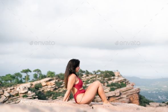 Caucasian mature woman sitting with her feet together barefoot in a swimsuit on a large rock in the