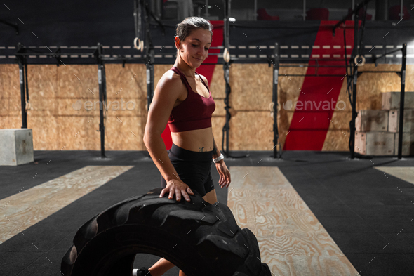 caucasian girl athlete standing smiling rolling a big crossfit tractor wheel in a big gym - Stock Photo - Images