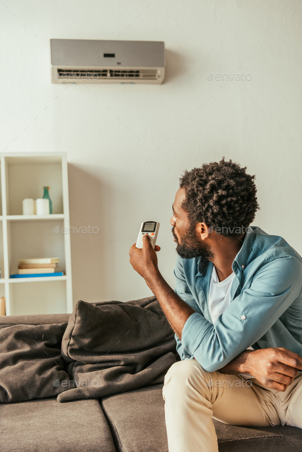 african american man holding air conditioner remote controller while sitting on couch at home