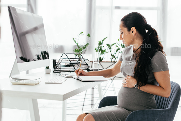 side view of pregnant woman sitting behind table with computer, keyboard and document tray, writing