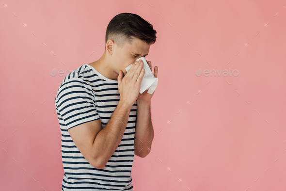 young man in striped t-shirt blowing nose and using napkin isolated on pink