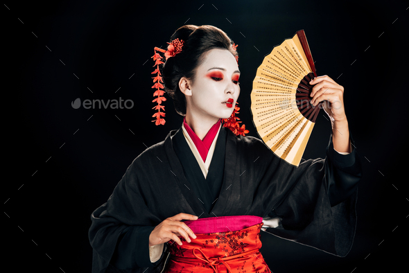geisha in kimono with red flowers in hair holding traditional asian hand fan isolated on black