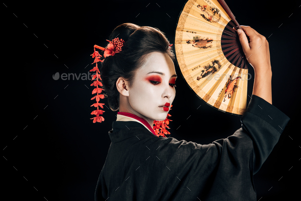 side view of geisha in black kimono with red flowers in hair holding traditional asian hand fan