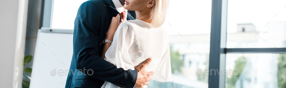 panoramic shot of businessman hugging blonde coworker while flirting in office