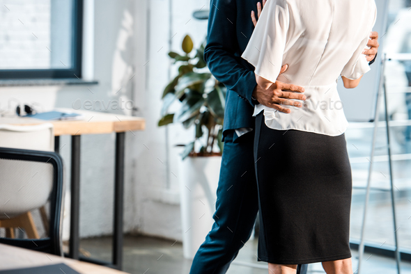 cropped view of businessman hugging businesswoman while standing and flirting in office