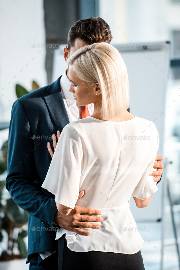 businessman hugging blonde and attractive coworker while flirting in office